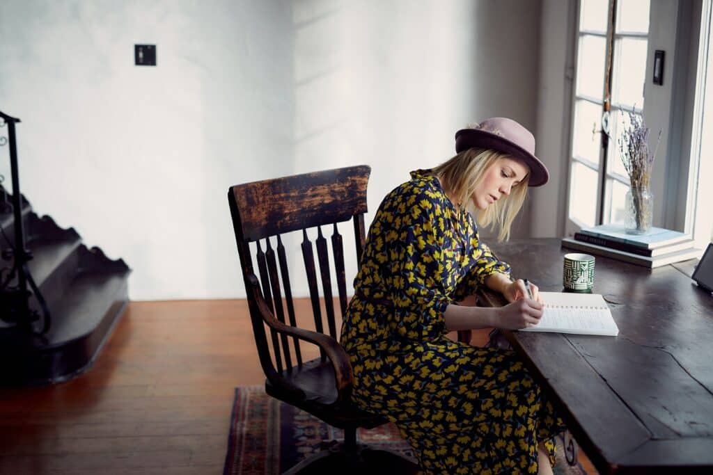 woman wearing a black/yellow floral dress and a hat writing on her desk